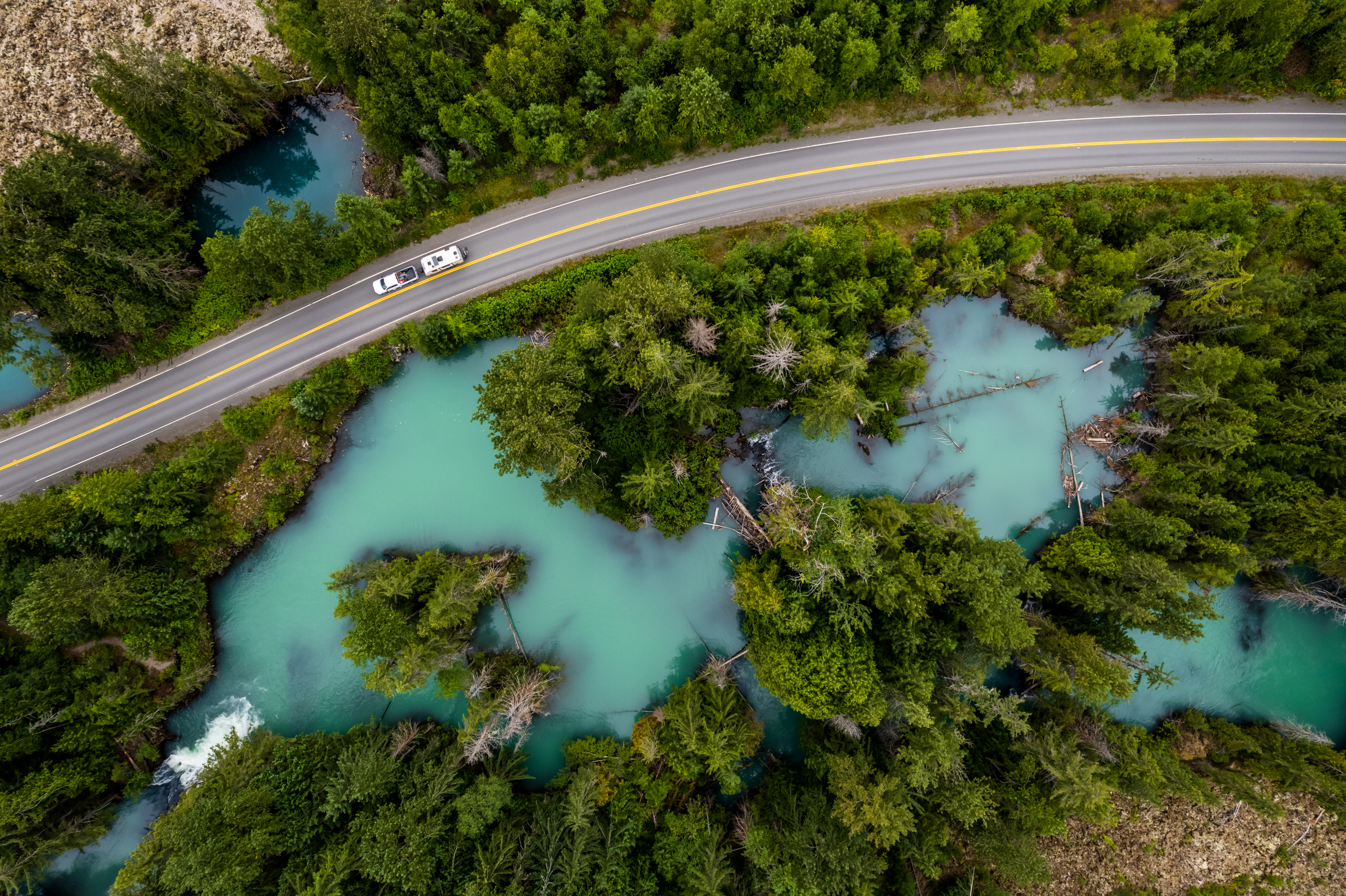 RV driving on a highway surrounded by glacier blue water and trees