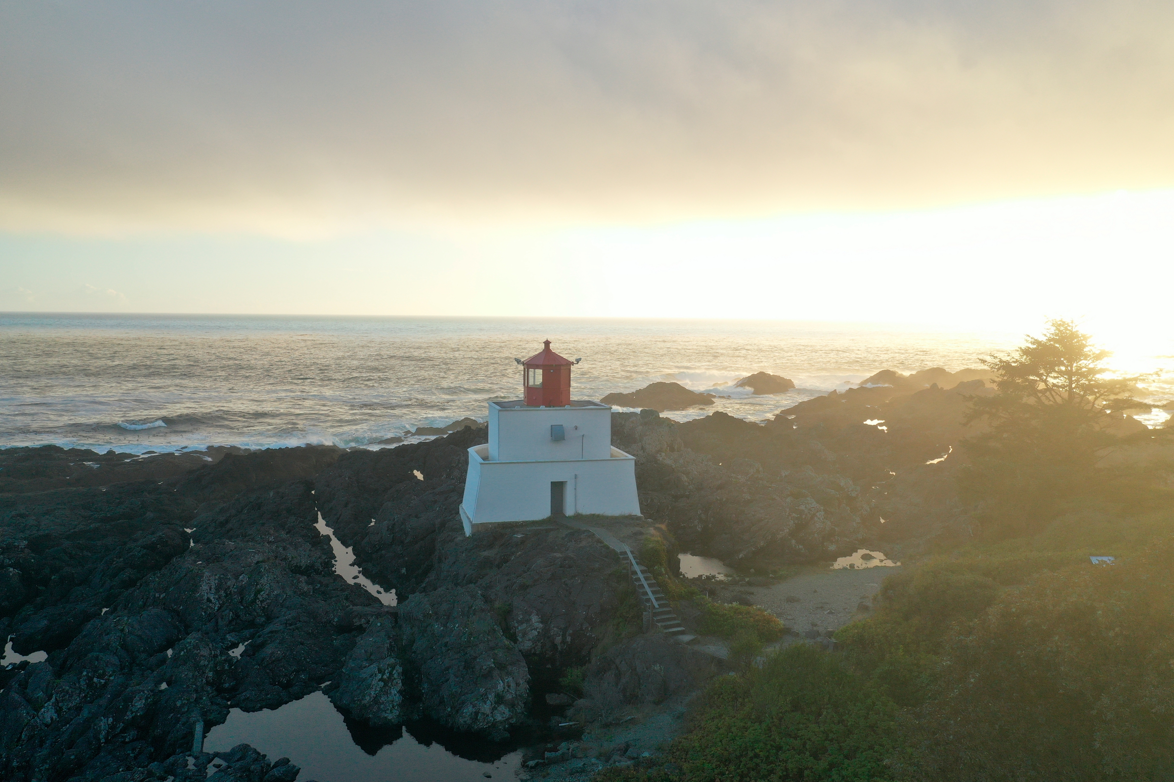 A short white building topped in red sits on a rocky coastline at sunset with the ocean in the background