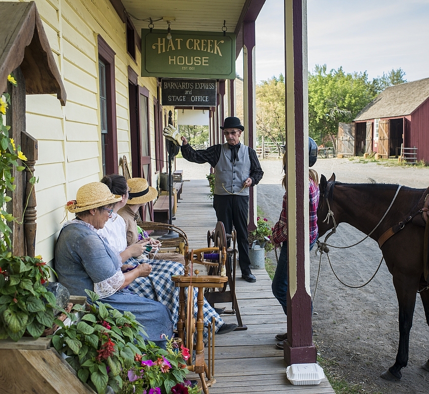 Actors in historical costume sit outside on a bench at Hat Creek Ranch. A horse is tethered to the building.