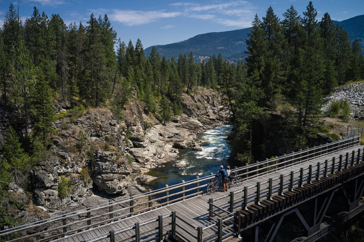 A couple has taken a break from biking on a trestle bridge, and is looking out at the canyon below.